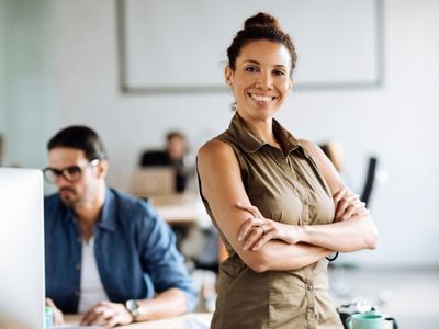 A smiling woman with her arms crossed stands in front of a desk with a man on a computer